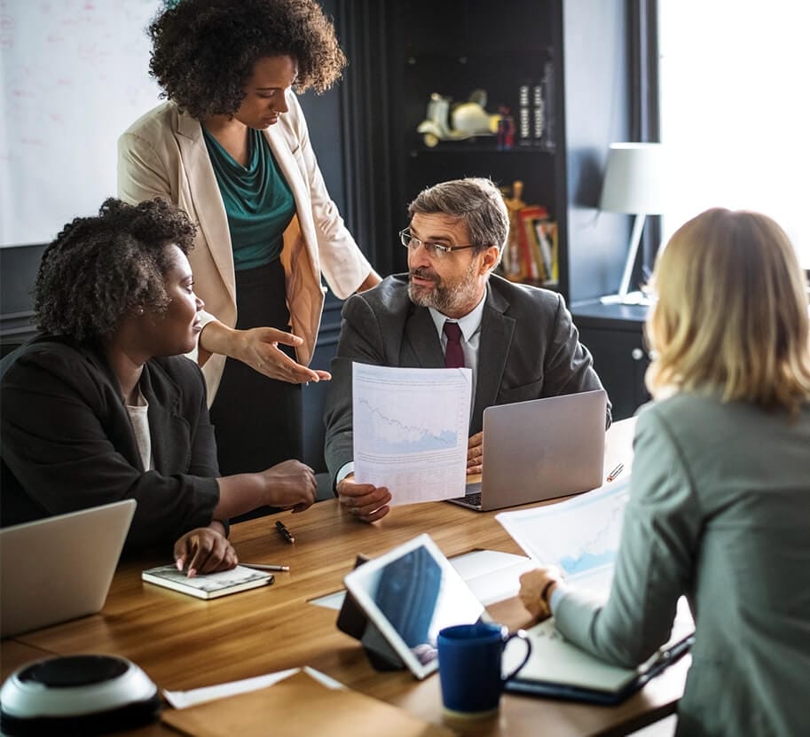 A diverse group of business professionals engaged in a discussion around a table with laptops, documents, and notepads.