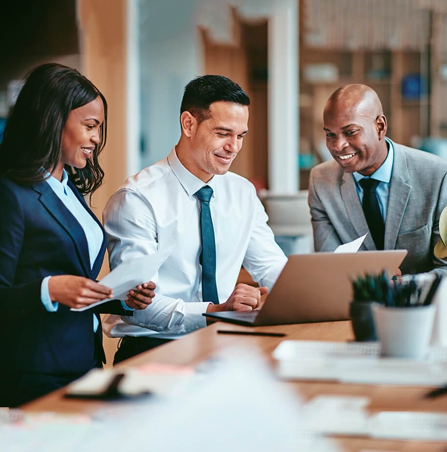 Three professionals collaborate around a laptop, discussing papers in a modern office setting.